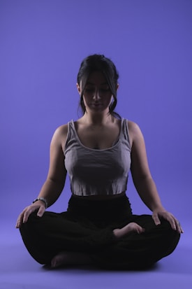 A person is sitting in a meditative pose with legs crossed on a smooth purple background. Their eyes appear closed, and they seem relaxed and focused. The lighting highlights the contours of their face and upper body, casting soft shadows.