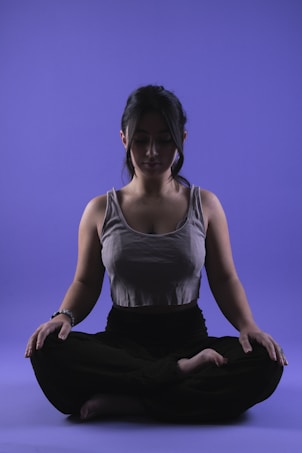 A person is sitting in a meditative pose with legs crossed on a smooth purple background. Their eyes appear closed, and they seem relaxed and focused. The lighting highlights the contours of their face and upper body, casting soft shadows.