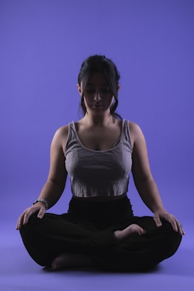 A person is sitting in a meditative pose with legs crossed on a smooth purple background. Their eyes appear closed, and they seem relaxed and focused. The lighting highlights the contours of their face and upper body, casting soft shadows.