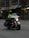 A police officer is riding a motorcycle with markings indicating it's from the Vancouver Police Department. The officer is wearing a helmet and a high-visibility jacket. The motorcycle has headlights on and features various lights on the front. The setting appears to be an urban street with buildings in the background.