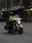 A police officer is riding a motorcycle with markings indicating it's from the Vancouver Police Department. The officer is wearing a helmet and a high-visibility jacket. The motorcycle has headlights on and features various lights on the front. The setting appears to be an urban street with buildings in the background.