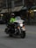 A police officer is riding a motorcycle with markings indicating it's from the Vancouver Police Department. The officer is wearing a helmet and a high-visibility jacket. The motorcycle has headlights on and features various lights on the front. The setting appears to be an urban street with buildings in the background.