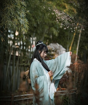 A person in traditional attire stands amidst a serene bamboo forest. They are wearing a flowing robe, gently holding a sleeve, and appear to be contemplating or absorbed in the natural surroundings. The scene is bathed in soft, muted light, creating a tranquil ambiance.
