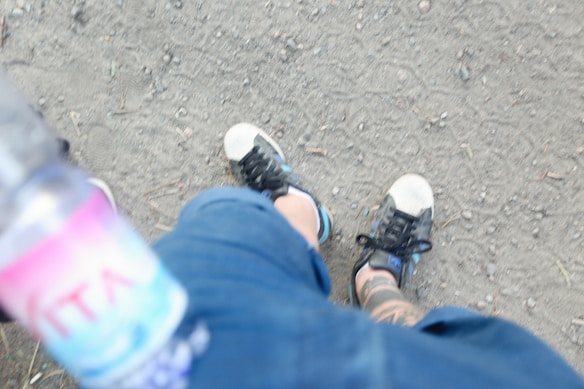 The image captures a pair of feet wearing black and white sneakers on a dirt path. The ground is slightly uneven with small stones scattered around. A tattoo is visible on the ankle of one leg, and a partially blurred water bottle is held in the foreground.