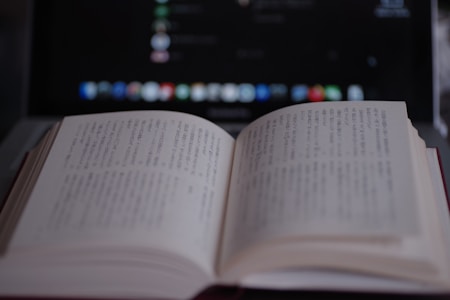 An open book with text in a non-Latin script is in focus, placed in front of a blurry, dark-screened laptop with visible icons and text.