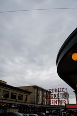 A cloudy sky looms over an urban market scene, featuring a prominent clock and neon sign reading 'PUBLIC MARKET CENTER'. The architecture is classic with several storefronts visible beneath the sign. People in the foreground add a sense of activity.