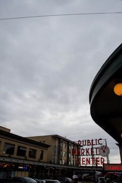 A cloudy sky looms over an urban market scene, featuring a prominent clock and neon sign reading 'PUBLIC MARKET CENTER'. The architecture is classic with several storefronts visible beneath the sign. People in the foreground add a sense of activity.