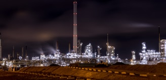 An industrial complex or refinery is lit up brightly against a dark night sky. Numerous lights illuminate various structures, towers, and machinery, some of which emit light trails or smoke clouds.