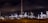 An industrial complex or refinery is lit up brightly against a dark night sky. Numerous lights illuminate various structures, towers, and machinery, some of which emit light trails or smoke clouds.