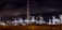 An industrial complex or refinery is lit up brightly against a dark night sky. Numerous lights illuminate various structures, towers, and machinery, some of which emit light trails or smoke clouds.