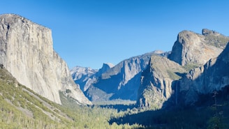A breathtaking view of a mountainous landscape with towering rock formations under a clear blue sky. The scene includes lush greenery in a valley and prominent cliffs rising majestically.