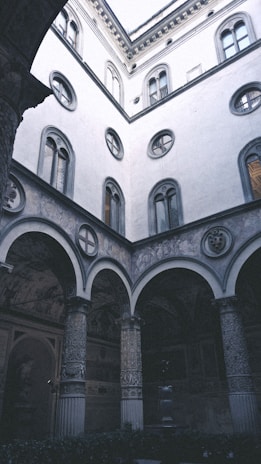 A picturesque courtyard of a historical building with ornate columns and arched windows. The walls feature intricate stonework and faded frescoes. The architecture reflects renaissance styles with circular and rectangular window designs and detailed carvings.