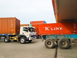 Several large shipping containers are being transported by trucks in a port area. The containers are primarily red and are being moved by vehicles that include a white Isuzu truck. The scene takes place in a spacious, open area with a clear sky.