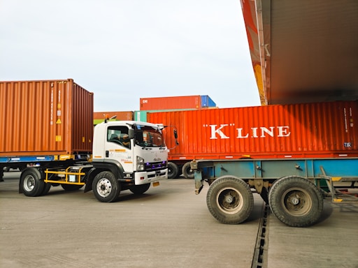 Several large shipping containers are being transported by trucks in a port area. The containers are primarily red and are being moved by vehicles that include a white Isuzu truck. The scene takes place in a spacious, open area with a clear sky.