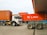 Several large shipping containers are being transported by trucks in a port area. The containers are primarily red and are being moved by vehicles that include a white Isuzu truck. The scene takes place in a spacious, open area with a clear sky.