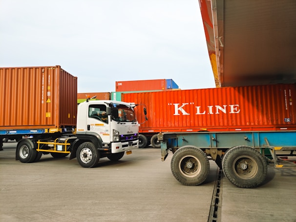 Several large shipping containers are being transported by trucks in a port area. The containers are primarily red and are being moved by vehicles that include a white Isuzu truck. The scene takes place in a spacious, open area with a clear sky.