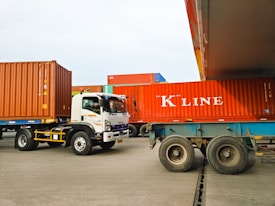 Several large shipping containers are being transported by trucks in a port area. The containers are primarily red and are being moved by vehicles that include a white Isuzu truck. The scene takes place in a spacious, open area with a clear sky.