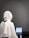 A person wearing a white hijab sits at a table working on a tablet. The background is a plain gray wall, and the focus is on the back of the person. The tablet displays text, suggesting focused work or study.