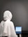 A person wearing a white hijab sits at a table working on a tablet. The background is a plain gray wall, and the focus is on the back of the person. The tablet displays text, suggesting focused work or study.