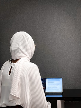 A person wearing a white hijab sits at a table working on a tablet. The background is a plain gray wall, and the focus is on the back of the person. The tablet displays text, suggesting focused work or study.