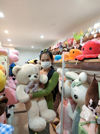 A person wearing a mask is holding a large white teddy bear in a store filled with various plush toys. Shelves around are packed with colorful stuffed animals, including characters with large eyes and small smiles. The lighting is bright and the scene appears cheerful.