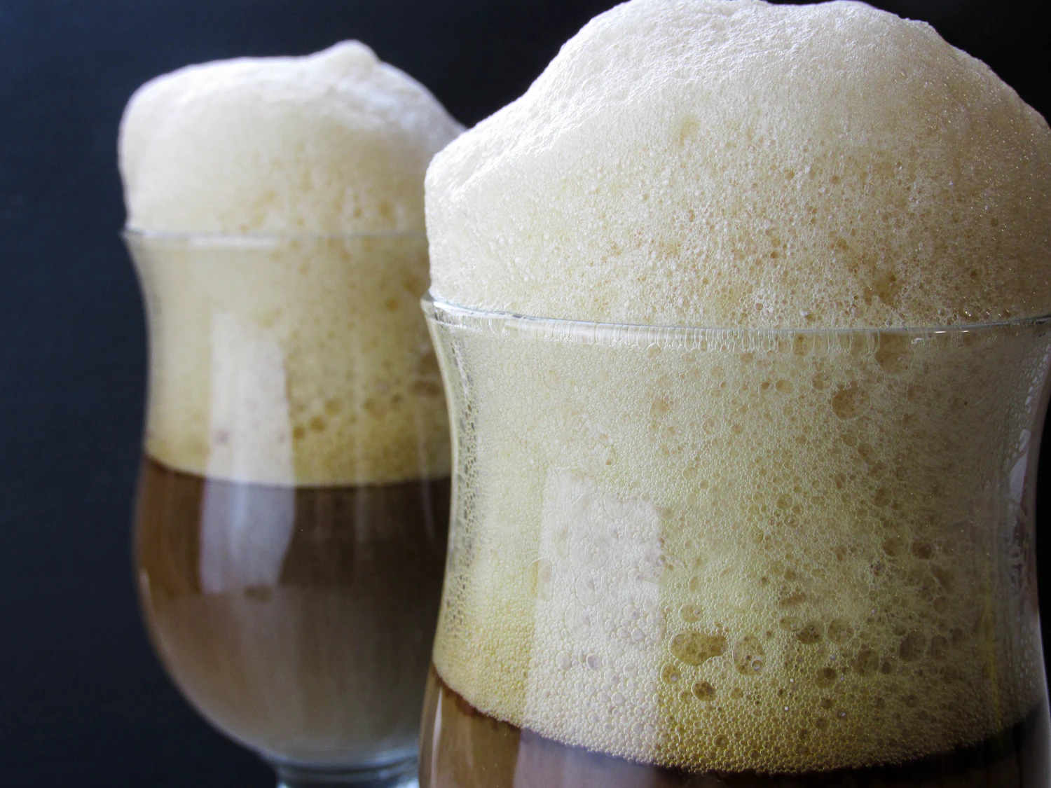 Two root-beer floats with tall heads of cream foam glowing amber against a dark booth