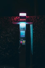A payphone stands illuminated by pink and blue lighting against a dark background. The payphone's handset is vibrant pink, and it reflects the neon glow. The surrounding area is dimly lit, adding a mysterious and nostalgic atmosphere.