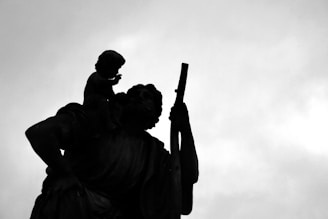 Silhouette of a statue featuring an adult figure holding a child and a staff against a cloudy sky.