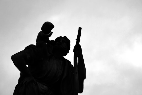 Silhouette of a statue featuring an adult figure holding a child and a staff against a cloudy sky.