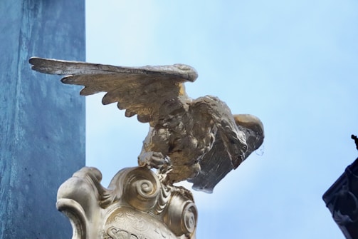 A close-up view of a gold-colored eagle statue perched atop an ornate pedestal. The wings appear to be semi-extended, and the eagle looks as if it is about to take flight. The background includes a clear blue sky and a structural element on the left side.