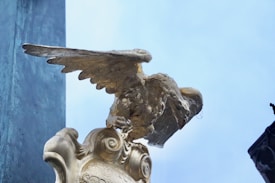 A close-up view of a gold-colored eagle statue perched atop an ornate pedestal. The wings appear to be semi-extended, and the eagle looks as if it is about to take flight. The background includes a clear blue sky and a structural element on the left side.