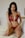 A person with long dark hair is wearing a red lace lingerie set. They are kneeling on a bed with light-colored sheets, and there are minimal decorations in the background.