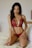 A person with long dark hair is wearing a red lace lingerie set. They are kneeling on a bed with light-colored sheets, and there are minimal decorations in the background.