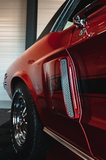 A close-up view of a red vintage car, focusing on the side panel and detailing. The shiny paint reflects light, highlighting a classic design with decorative elements such as a chrome air vent and a stripe with the letters 'GCS'. The wheel is visible with polished chrome rims, giving a sense of classic automotive craftsmanship.