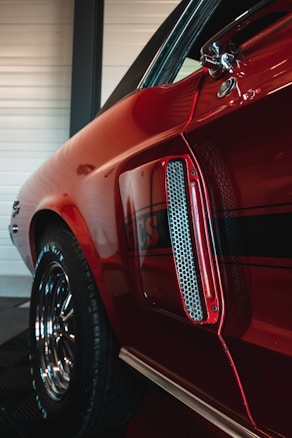 A close-up view of a red vintage car, focusing on the side panel and detailing. The shiny paint reflects light, highlighting a classic design with decorative elements such as a chrome air vent and a stripe with the letters 'GCS'. The wheel is visible with polished chrome rims, giving a sense of classic automotive craftsmanship.