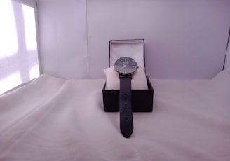 A wristwatch is displayed in an open black box, resting on a white cushion. The watch features a sleek black strap and a classic round watch face.