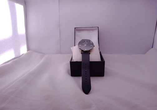 A wristwatch is displayed in an open black box, resting on a white cushion. The watch features a sleek black strap and a classic round watch face.