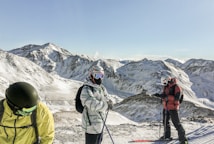 Three people dressed in ski gear are standing on a snowy mountain slope. The backdrop consists of rugged, snow-covered mountains and a clear blue sky. The individuals are wearing ski helmets and goggles, with ski poles in their hands. Their clothing includes colorful jackets, and they appear to be preparing for a ski descent.