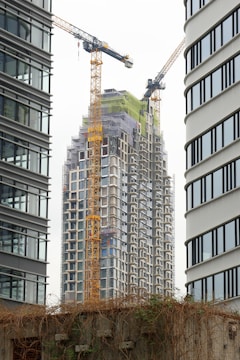 A high-rise building under construction is framed between two modern office towers. Two large yellow cranes are actively involved in the construction process. The building is covered in scaffolding, indicating ongoing work. A concrete wall with climbing plants is visible in the foreground.