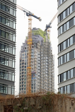 A high-rise building under construction is framed between two modern office towers. Two large yellow cranes are actively involved in the construction process. The building is covered in scaffolding, indicating ongoing work. A concrete wall with climbing plants is visible in the foreground.