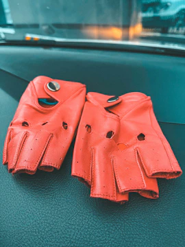 A pair of bright orange leather driving gloves with button closures is resting on a dark textured surface, likely a car dashboard. The gloves have several small perforations and cut-out sections on the knuckles for better flexibility and breathability.
