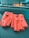 A pair of bright orange leather driving gloves with button closures is resting on a dark textured surface, likely a car dashboard. The gloves have several small perforations and cut-out sections on the knuckles for better flexibility and breathability.