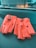 A pair of bright orange leather driving gloves with button closures is resting on a dark textured surface, likely a car dashboard. The gloves have several small perforations and cut-out sections on the knuckles for better flexibility and breathability.