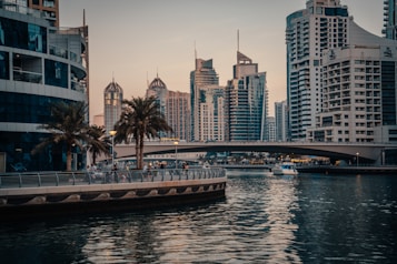 A vibrant cityscape featuring modern skyscrapers with unique architectural designs. A bridge spans across a tranquil body of water, over which a small boat sails. Palm trees line the waterfront promenade where people are walking and enjoying the outdoor space. The scene is captured during a calm evening with a soft, warm light.
