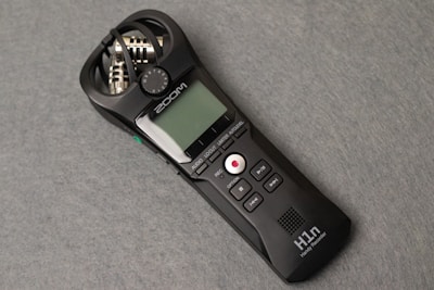 A sleek, handheld audio recorder in black with a small display screen, various control buttons, and built-in microphones. The design includes a visible brand name, and the device rests on a flat grey surface.
