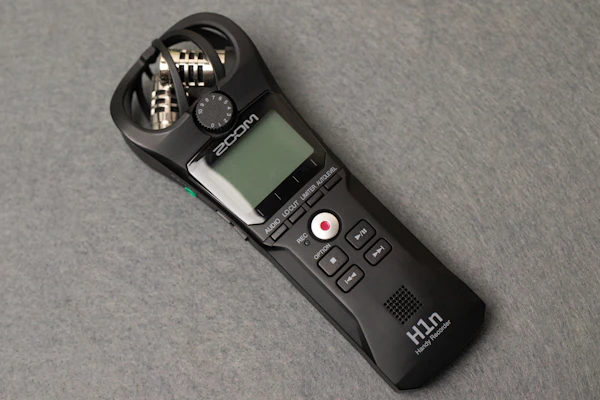 A sleek, handheld audio recorder in black with a small display screen, various control buttons, and built-in microphones. The design includes a visible brand name, and the device rests on a flat grey surface.