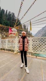 A person stands on a patterned metal platform with hands in pockets, surrounded by colorful string lights and flags. A temple is visible in the background, nestled among tall evergreen trees in a mountainous area.