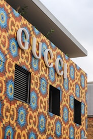 A colorful, patterned wall featuring the Gucci logo in large, bold white letters. The wall displays a vibrant design with repeating blue and yellow motifs set against a brick background. There are small windows and vents embedded within the wall.