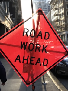 A bright orange road work sign with the words 'ROAD WORK AHEAD' stands prominently on a busy street. The sun casts a glare through the sign, illuminating it. In the background, urban buildings and cars fill the street, while a person is partially visible on the left side of the frame.