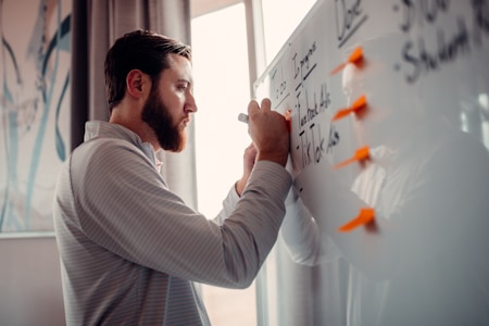 A man is writing on a whiteboard with a marker. The whiteboard contains a list and has orange sticky notes attached to it. The man appears focused and is wearing a striped shirt. Soft lighting from a window illuminates the scene.